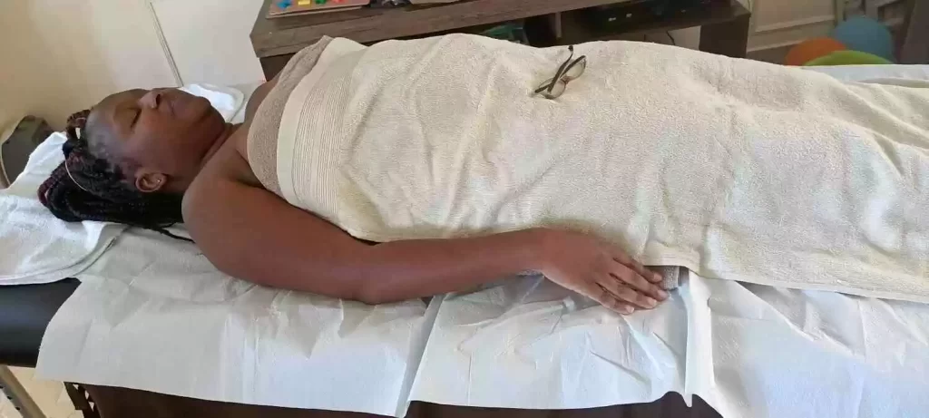 Client, a lady lying on her back on a massage table, covered in a towel, during a full-body massage treatment in her home in Nairobi
